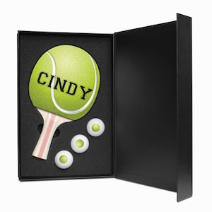 May include: A black gift box with a white and pink tennis paddle with the name "Cindy" on the paddle face. The box is open and there are three white tennis balls inside.