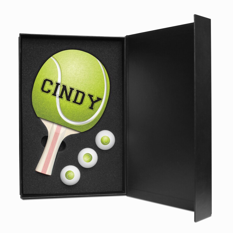 May include: A black gift box with a white and pink tennis paddle with the name "Cindy" on the paddle face. The box is open and there are three white tennis balls inside.