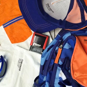 May include: A blue and orange camouflage backpack with a zippered pocket and an orange pull tab. The backpack has a white baseball inside. There is a blue baseball cap with an orange brim, a white t-shirt with orange sleeves, a pair of blue sunglasses, and a red stamp with the word "Trodat" on it.