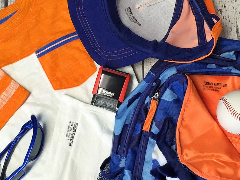 May include: A blue and orange camouflage backpack with a zippered pocket and an orange pull tab. The backpack has a white baseball inside. There is a blue baseball cap with an orange brim, a white t-shirt with orange sleeves, a pair of blue sunglasses, and a red stamp with the word "Trodat" on it.