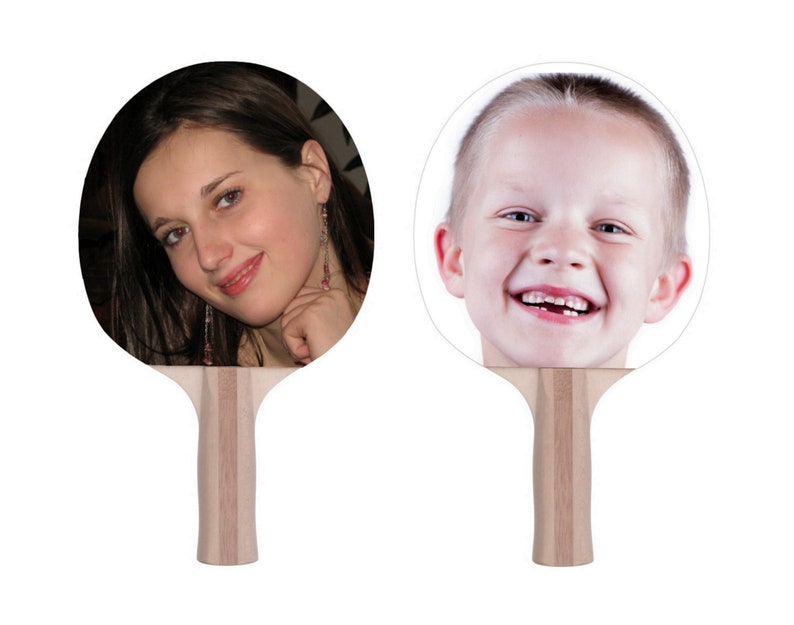 May include: Two white ping pong paddles with a photo of a person's face on each side. The paddle on the left has a photo of a woman with long brown hair. The paddle on the right has a photo of a young boy with short blonde hair.
