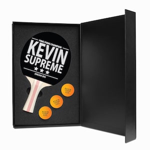 May include: A black ping pong paddle with the text "KEVIN SUPREME" and three stars, along with three orange ping pong balls, all presented in a black gift box. The paddle also has the text "PING PONG CHAMPION" and "DREAM BIG".
