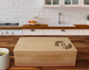 May include: A natural bamboo tea box with a hinged lid. The lid is engraved with a floral wreath design and the letter "B".