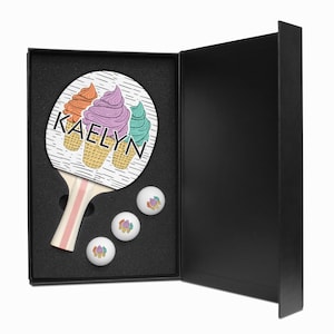 May include: A black gift box with a white and pink ping pong paddle with a design of three ice cream cones and the name "KAELYN" on it. The box is open and three white ping pong balls with ice cream cone designs are visible.