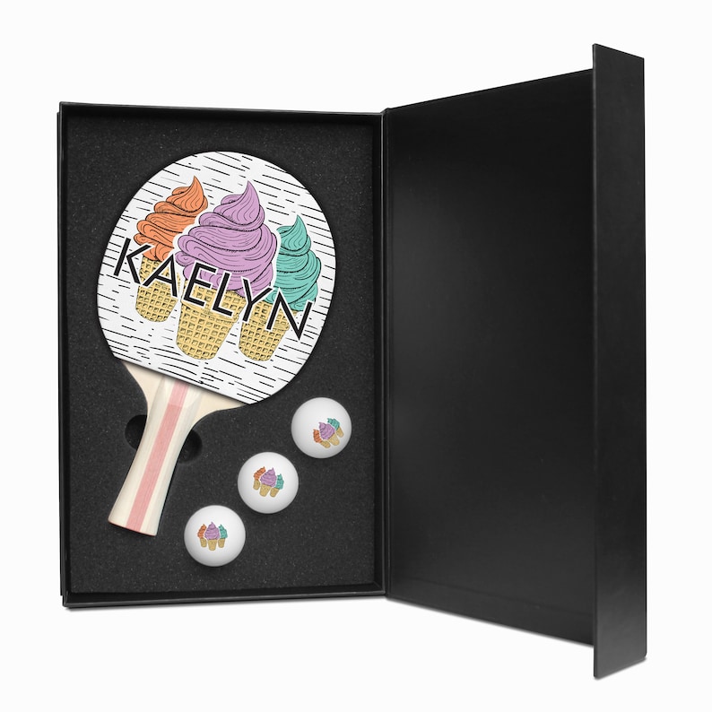 May include: A black gift box with a white and pink ping pong paddle with a design of three ice cream cones and the name "KAELYN" on it. The box is open and three white ping pong balls with ice cream cone designs are visible.