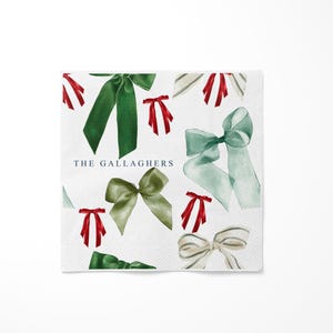 May include: White square napkin with a pattern of watercolor bows in shades of green, red, and gray. The text "THE GALLAGHERS" is printed in a dark blue font. The bows are of varying sizes and styles.