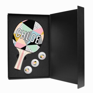 May include: A black box with a pink, blue, and yellow geometric patterned ping pong paddle with the name "CHLOE" printed on it. The box is open and three white ping pong balls are visible inside.