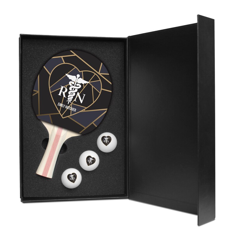 May include: A black and gold ping pong paddle set with a heart design and the text "RN" and "Emily Hatcher". The paddle is in a black box with three white ping pong balls.