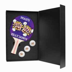 May include: A purple and yellow ping pong paddle with the text "Noah's Beat Down" and a graphic of fists with the words "Ring Ring" on them. The paddle is in a black box with three white ping pong balls with the word "POW" on them.