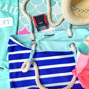 May include: A blue and white striped tote bag with a rope handle. The bag has a light blue pocket with the name "Brianna Murphy" printed on it. There is a pink and orange striped towel with the same name printed on it. A blue shirt with the text "Mermaid Academy" is also visible.