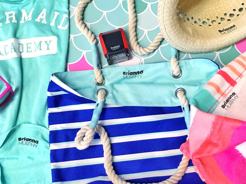 May include: A blue and white striped tote bag with a rope handle. The bag has a light blue pocket with the name "Brianna Murphy" printed on it. There is a pink and orange striped towel with the same name printed on it. A blue shirt with the text "Mermaid Academy" is also visible.