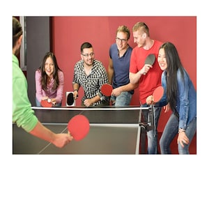 May include: A group of people playing table tennis. They are using red and black paddles and a white ball. The table is black and has a net in the middle. The people are smiling and having fun.
