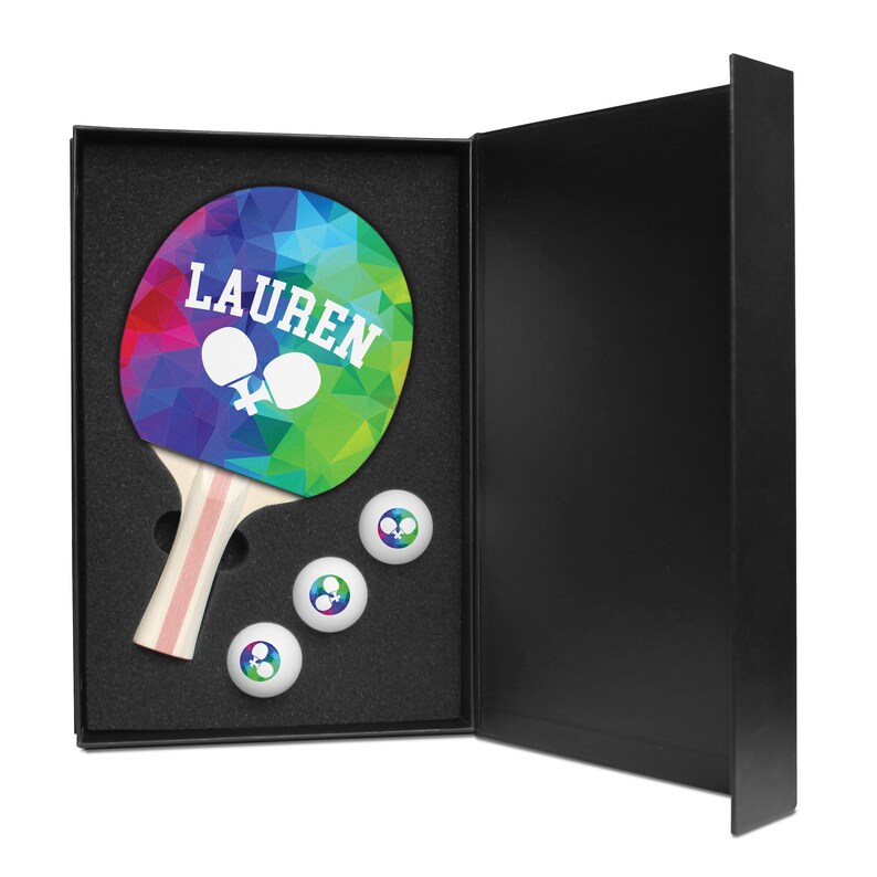 May include: A black gift box with a rainbow-colored ping pong paddle with the name "LAUREN" printed on it. The box also contains three white ping pong balls.