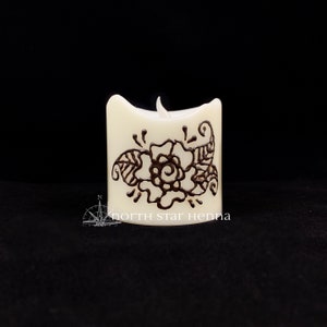 Peut inclure: Une bougie blanche avec un motif de henné brun représentant une fleur et des feuilles. La bougie est allumée et la flamme est visible.