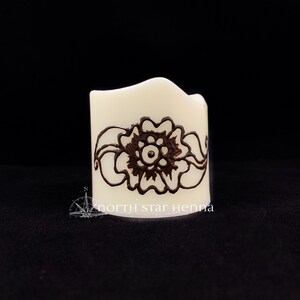 Peut inclure: Une bougie blanche avec un motif de henné brun représentant une fleur et des feuilles. La bougie porte l'inscription "north star henna" sur le fond.