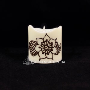 Peut inclure: Une bougie blanche avec un motif de henné brun en forme de fleur. La bougie a une mèche et est allumée. Le texte "north star henna" est imprimé au bas de la bougie.
