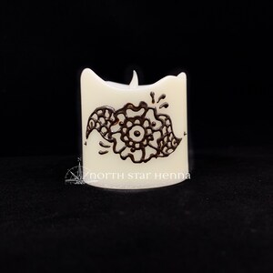 May include: White candle with a brown henna design on the side. The design is a stylized floral pattern. The candle is lit and the flame is visible. The text "north star henna" is printed on the bottom of the candle.