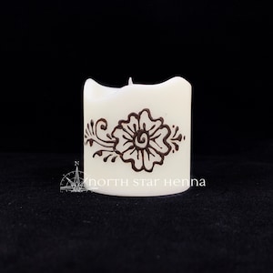 Peut inclure: Bougie blanche avec un motif au henné brun représentant une fleur et des feuilles. La bougie porte l'inscription "north star henna" sur le côté.