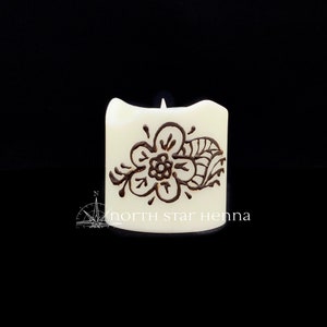 Peut inclure: Bougie blanche avec un motif au henné brun représentant une fleur et des feuilles. La bougie porte l'inscription "north star henna" sur le fond.