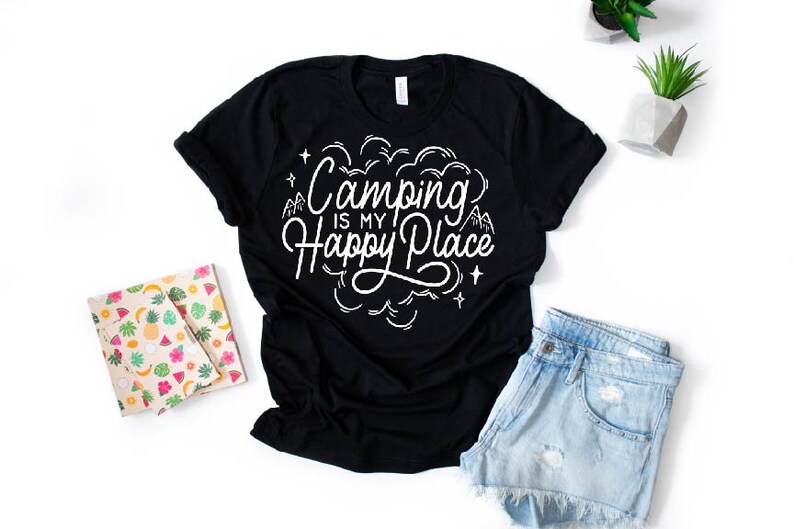 Może przedstawiać: Czarny T-shirt z białym napisem "Camping is my Happy Place". Tekst jest w stylizowanym foncie i zawiera małe rysunki g&oacute;r i gwiazd.