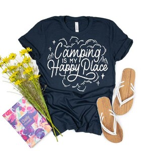 Może przedstawiać: Koszulka w kolorze granatowym z białym napisem "Camping is my happy place". Koszulka leży na białym tle z bukietem ż&oacute;łtych kwiat&oacute;w i parą brązowo-białych klapek.