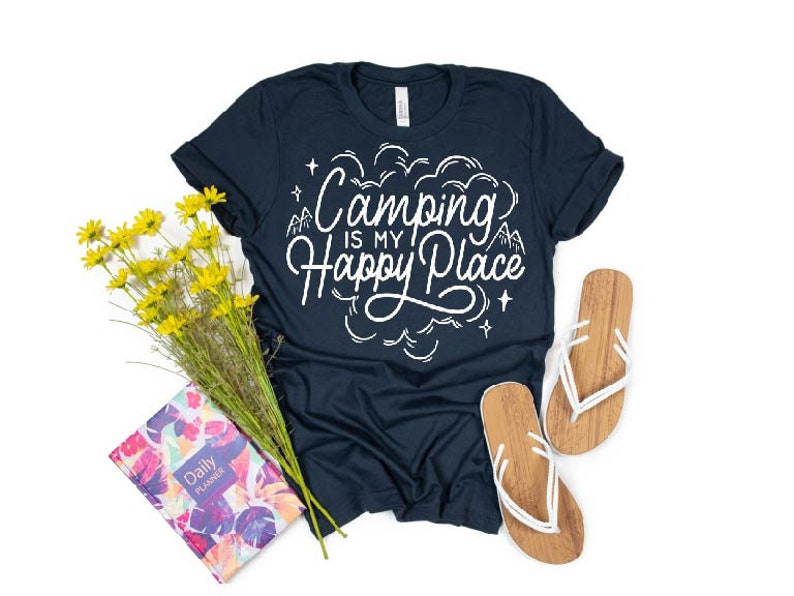 Może przedstawiać: Koszulka w kolorze granatowym z białym napisem "Camping is my happy place". Koszulka leży na białym tle z bukietem ż&oacute;łtych kwiat&oacute;w i parą brązowo-białych klapek.