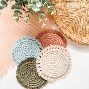 May include: A set of four circular coasters crafted from knotted cord in light blue, terracotta, olive green, and cream. Each coaster features a spiral pattern radiating from the center, finished with a short, fringed edge around the entire perimeter.