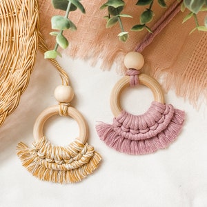 May include: Two macrame car air fresheners. One is mustard yellow and the other is dusty rose. Each has a wooden ring and bead, with a knotted fringe design. The air fresheners hang from a matching cord.