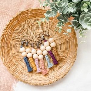 May include: A collection of handmade keychains with wooden beads and colorful macrame accents. Each keychain features a different color of braided macrame, including blue, yellow, pink, and purple. The keychains are displayed in a woven basket.