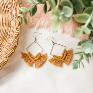 May include: A pair of gold-colored, geometric earrings with a mustard-yellow macrame design. The earrings have a square frame with a fringe of knotted threads. The earrings are displayed on a white surface.