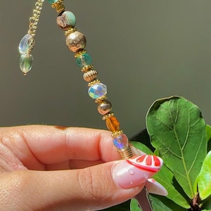 May include: A decorative hair accessory featuring a gold chain and an array of colourful beads. The beads are iridescent, gold, brown, and teal. The accessory is held in a hand with a red and white design on the fingernail.