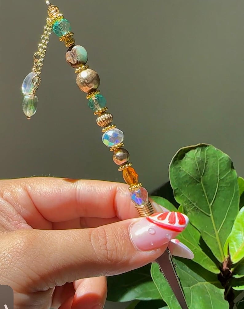 May include: A decorative hair accessory featuring a gold chain and an array of colourful beads. The beads are iridescent, gold, brown, and teal. The accessory is held in a hand with a red and white design on the fingernail.