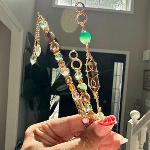 May include: Three handmade hair accessories with gold wire and colourful beads. One features a chain of clear and green beads, another has a green orb, and the third has a brown crystal. The accessories are held up in front of a window.