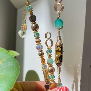 May include: Three handmade wire-wrapped crystal and bead pendants. The pendants feature gold-coloured wire, various beads in shades of green, gold, and iridescent, and a heart-shaped charm. One pendant has a tiger's eye crystal.