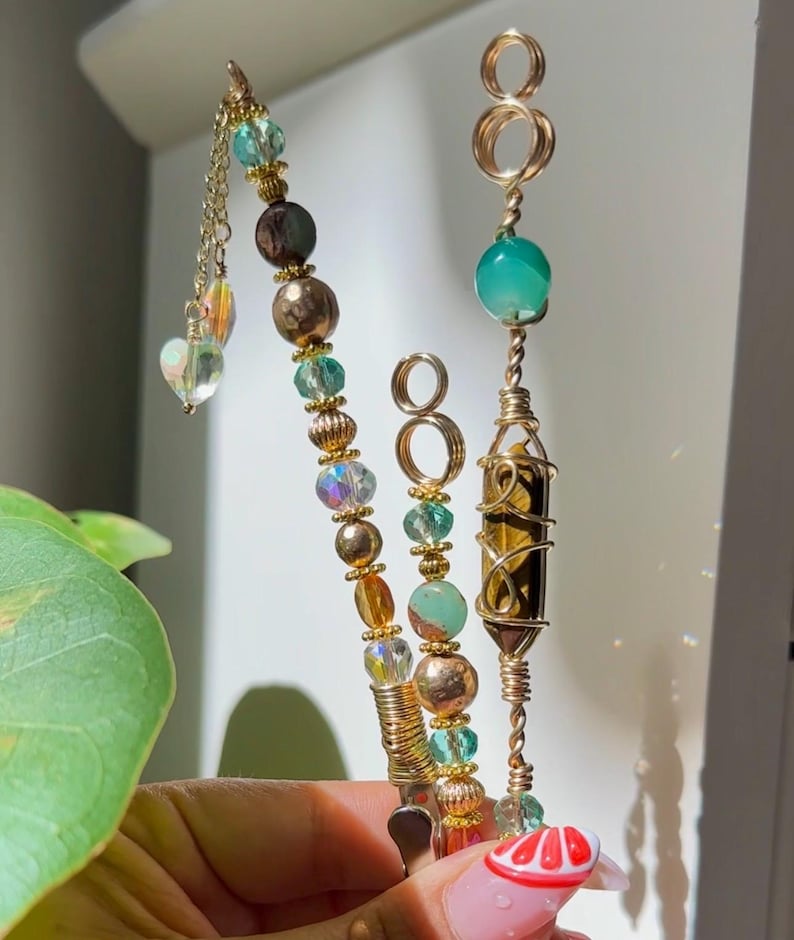 May include: Three handmade wire-wrapped crystal and bead pendants. The pendants feature gold-coloured wire, various beads in shades of green, gold, and iridescent, and a heart-shaped charm. One pendant has a tiger's eye crystal.