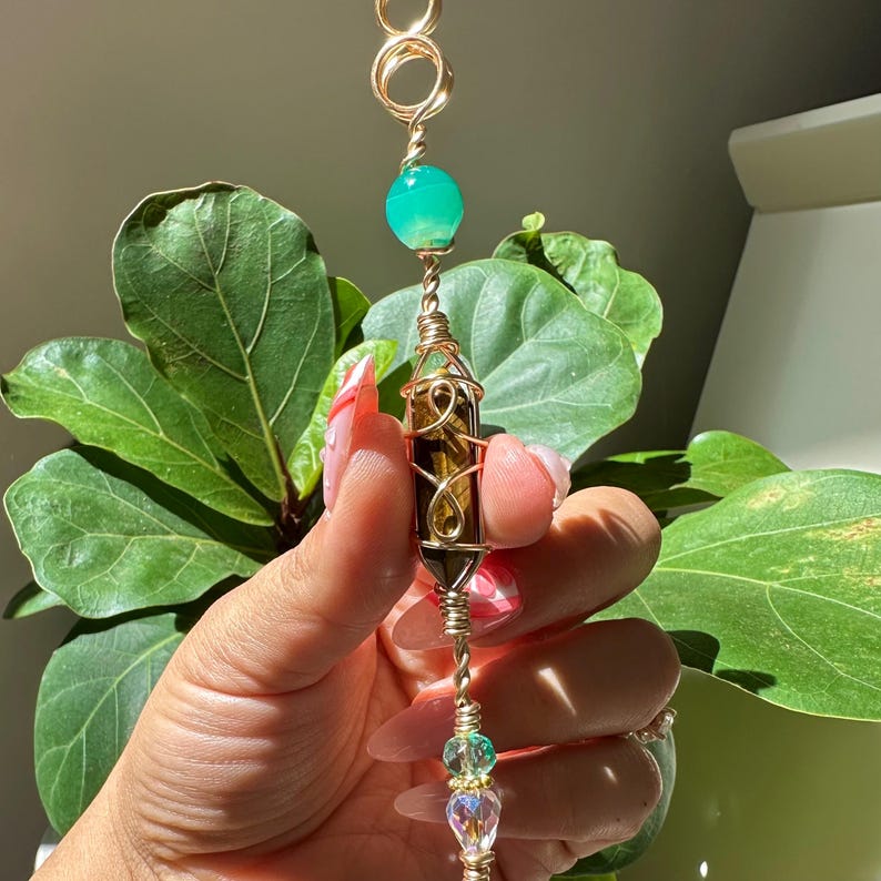 May include: A gold-coloured wire-wrapped crystal pendant. It features a tiger's eye crystal, a green sphere, and a clear crystal with iridescent accents. The design includes intricate wire wrapping and decorative beads, suitable for jewellery.
