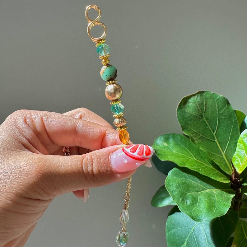 May include: A gold-coloured hair accessory featuring a sequence of beads in green, gold, and orange hues. The accessory has a loop at the top and a dangling chain with a clear crystal. The accessory is being held.