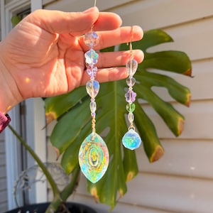 Puede incluir: Dos atrapasoles de cristal con alambre dorado y cuentas transparentes. Uno tiene un cristal de forma ovalada con patrones iridiscentes coloridos. El otro tiene un cristal redondo en la parte inferior y cuentas en forma de flor. Los atrapasoles cuelgan frente a una hoja verde.