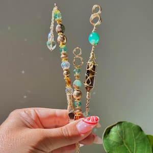 May include: Three handmade smoking accessories with gold wire, beads, and crystals. One features a pointed shaped crystal, another has a green stone, and the third has a tiger eye. The accessories are held in a hand.