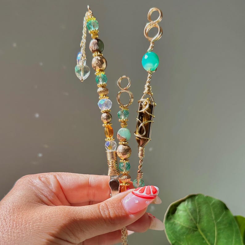 May include: Three handmade smoking accessories with gold wire, beads, and crystals. One features a pointed shaped crystal, another has a green stone, and the third has a tiger eye. The accessories are held in a hand.