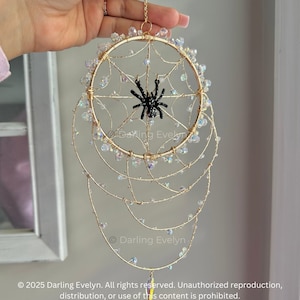 May include: A gold wire dream catcher with a black spider in the centre. The dream catcher has iridescent beads and a feather hanging from the bottom.