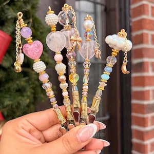 Puede incluir: Una mano sostiene cinco clips decorativos dorados, cada uno adornado con dijes de cuentas únicos. Los dijes incluyen corazones rosas, perlas, cristales transparentes y una nube con una luna creciente. Los clips están adornados con alambre dorado.