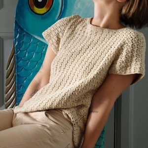 Op de afbeelding: Een beige gehaakte top met korte mouwen en een textuurpatroon. De top wordt gecombineerd met een beige broek. De persoon zit op een turquoise stoel met een visontwerp.