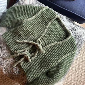 May include: Close-up of a sage green knitted top with a square neckline and a tie closure. The top is made of a ribbed knit fabric. The top is lying on a gray, textured surface.