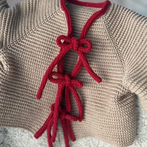 May include: A tan-colored, knitted cardigan with a red trim and tie closure. The cardigan features a textured knit pattern and a decorative red tie closure that adds a pop of color and visual interest to the garment.