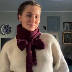 Peut inclure: Écharpe tricotée bordeaux avec un détail en forme de nœud. L'écharpe est portée sur un pull crème. La couleur de l'écharpe contraste avec le pull, créant un accessoire visuellement attrayant. Un ajout élégant à toute tenue.