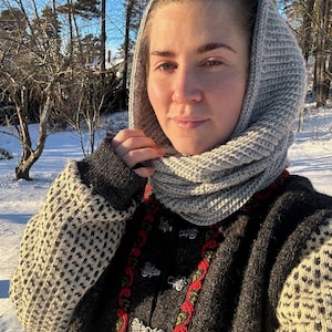 Può includere: Una persona indossa un cappuccio di maglia grigio e un maglione grigio scuro con un motivo. Il maglione presenta dettagli ricamati rossi e verdi e chiusure argentate. Lo sfondo mostra un paesaggio innevato con alberi.