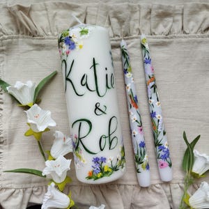 May include: White pillar candle and two taper candles, all adorned with hand-painted floral designs in shades of blue, purple, and yellow. The pillar candle features the names "Katie & Rob" in elegant script. Decorative candles for special occasions.