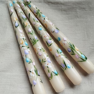 Może przedstawiać: Cztery kremowe świece stożkowe z ręcznie malowanymi wzorami kwiatowymi. Świece mają białe kwiaty, zielone liście i niebieskie i fioletowe akcenty.