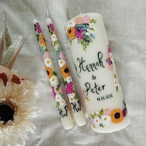 Könnte beinhalten: Ein Set aus drei elfenbeinfarbenen Kerzen, zwei Stabkerzen und einer Säulenkerze, verziert mit handgemalten Blumenmustern in Rosa, Orange, Gelb und Weiß. Die Säulenkerze ist mit den Namen "Hannah & Peter" und dem Datum "11.06.2026" beschriftet.